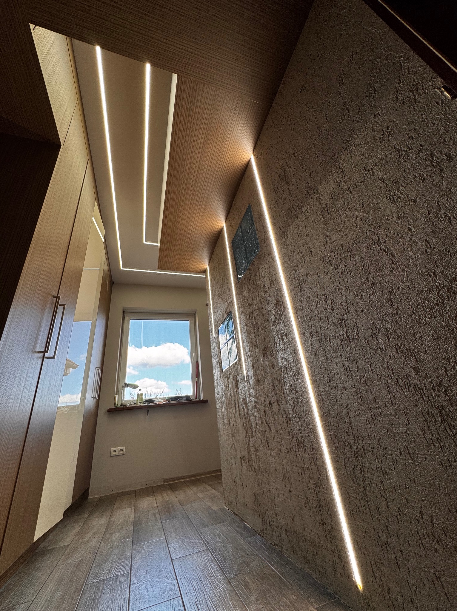 Wnętrze korytarza z nowoczesnym oświetleniem liniowym LED w suficie i ścianie, drewniana podłoga i szafa w zabudowie, okno z widokiem na niebo.