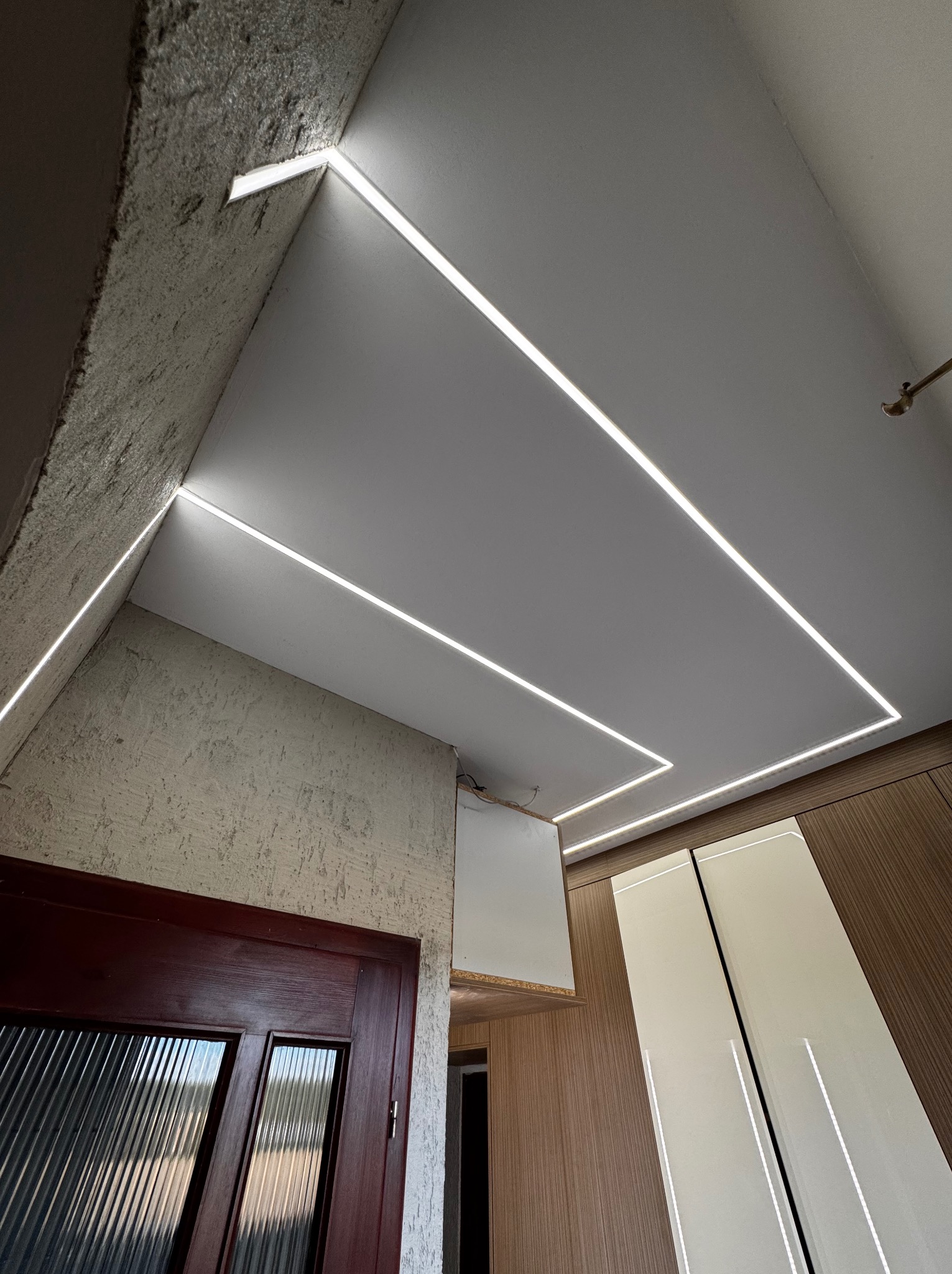 Nowoczesne oświetlenie sufitowe LED w kształcie geometrycznym, z widoczną surową fakturą ściany i fragmentem drzwi wejściowych