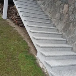 Zewnętrzne schody z szarego granitu prowadzące do domu z elewacją z kamienia naturalnego, obok ułożone drewno kominkowe.