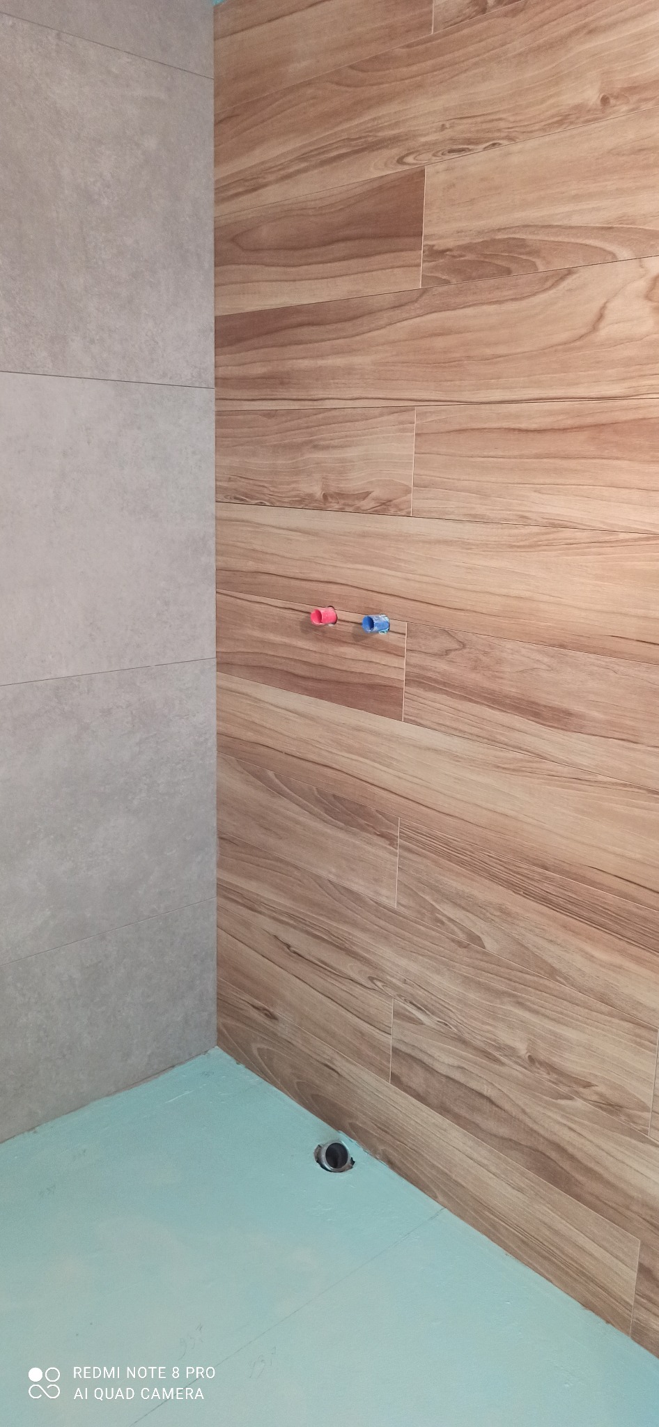 Ściana łazienki wykończona płytkami imitującymi drewno, z widocznymi przyłączami wodnymi z czerwonym i niebieskim korkiem oraz fragmentem ściany z szarymi płytkami, a także przygotowane podłoże...