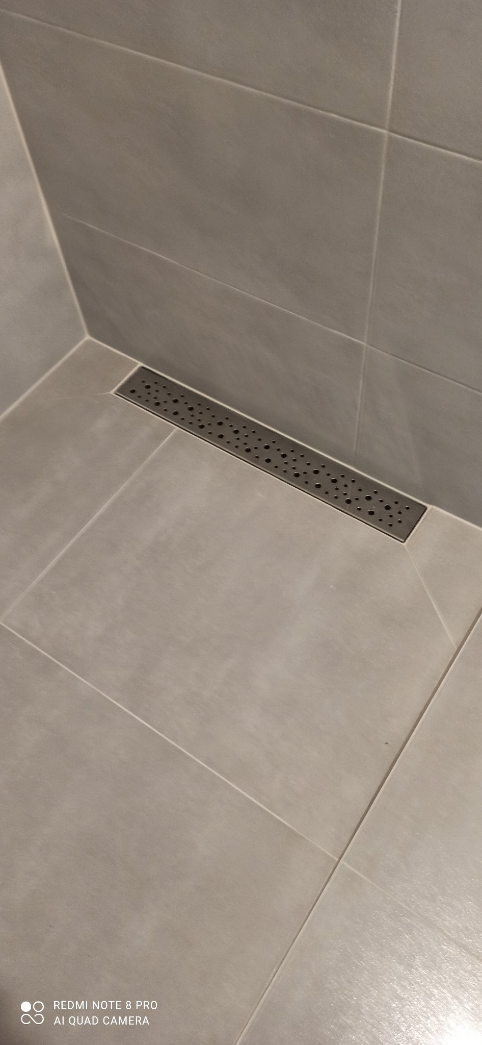 Szare płytki podłogowe i ścienne w kabinie prysznicowej z liniowym odpływem podłogowym z perforowaną maskownicą.
