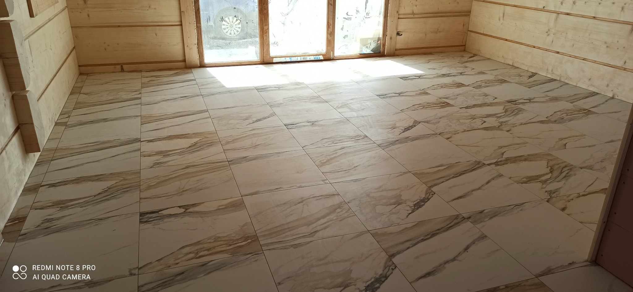 Ułożone marmurowe płytki podłogowe w jasnym, surowym wnętrzu z drewnianymi ścianami i oknem w tle.