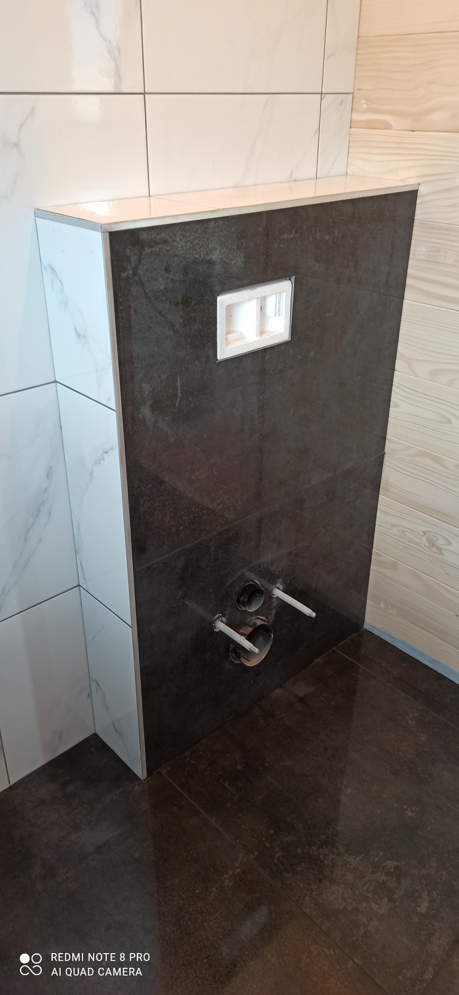 Częściowo obudowany stelaż podtynkowy WC z widocznymi rurami instalacyjnymi, obłożony ciemnymi płytkami imitującymi kamień, sąsiadujący z białymi płytkami marmurkowymi i drewnianą boazerią.