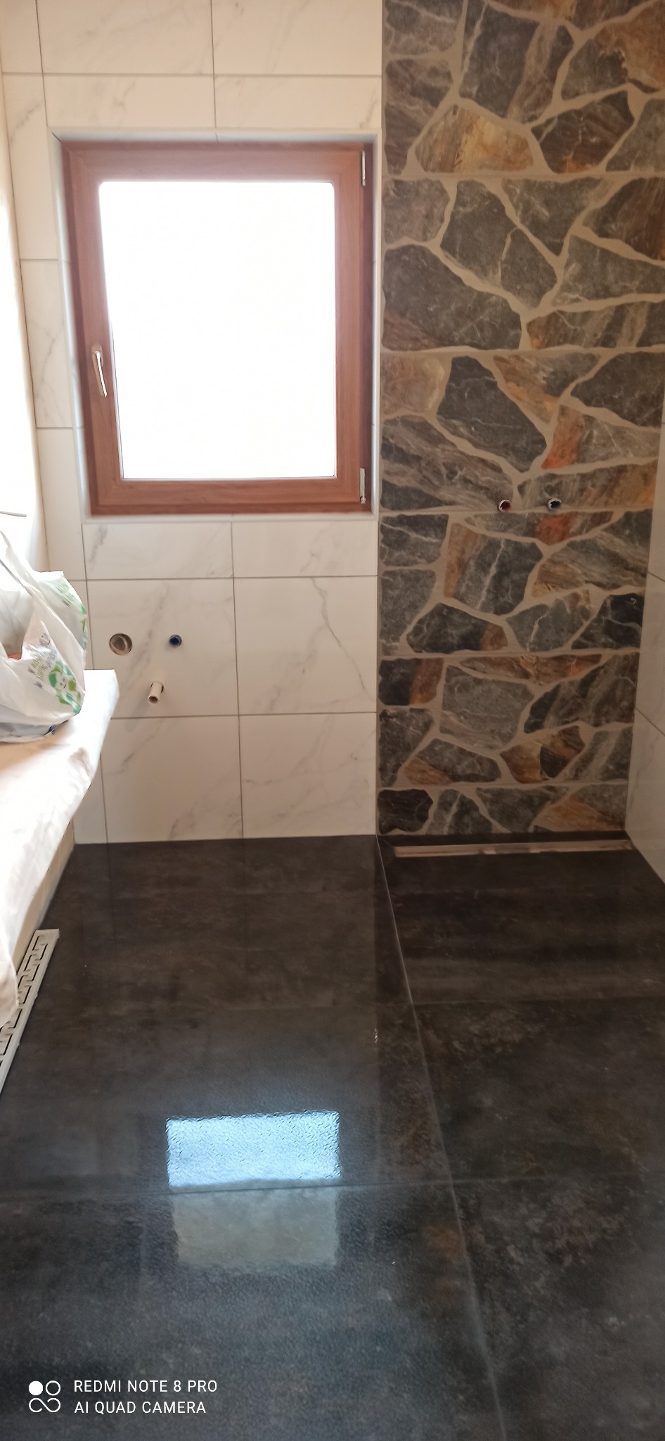 Wnętrze łazienki w trakcie wykańczania: ściana z kamienną mozaiką, białe płytki imitujące marmur, ciemna, błyszcząca podłoga z odpływem liniowym i drewniane obramowanie okna.