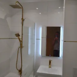 Nowoczesna łazienka z białymi płytkami, złotą armaturą i kabiną prysznicową z szybą. W lustrze odbija się osoba robiąca zdjęcie telefonem.