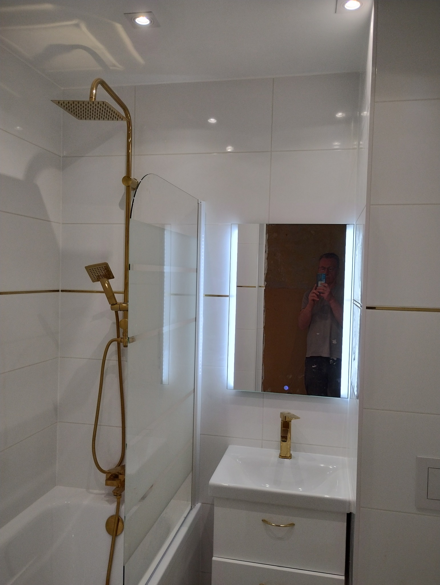 Nowoczesna łazienka z białymi płytkami, złotą armaturą i kabiną prysznicową z szybą. W lustrze odbija się osoba robiąca zdjęcie telefonem.
