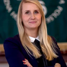 Portret uśmiechniętej blondwłosej kobiety w granatowej marynarce z rękami skrzyżowanymi na tle zielonego banera z logo, Sokółka.