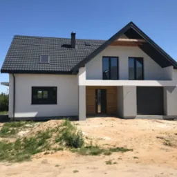 Nowoczesny dom jednorodzinny z grafitowym dachem i białymi ścianami, widok od frontu, teren wokół domu w trakcie zagospodarowywania.