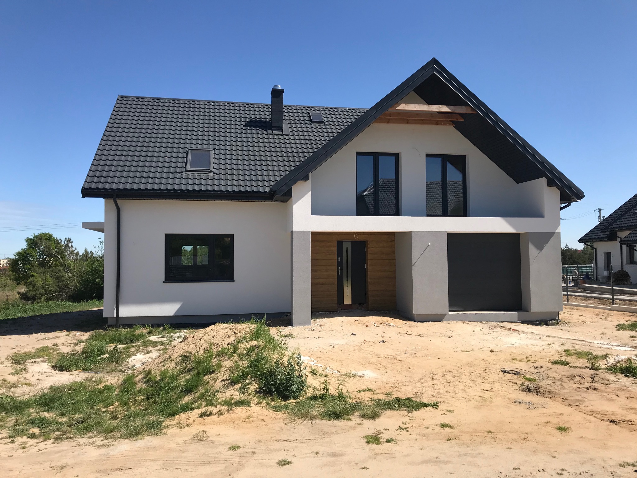 Nowoczesny dom jednorodzinny z grafitowym dachem i białymi ścianami, widok od frontu, teren wokół domu w trakcie zagospodarowywania.