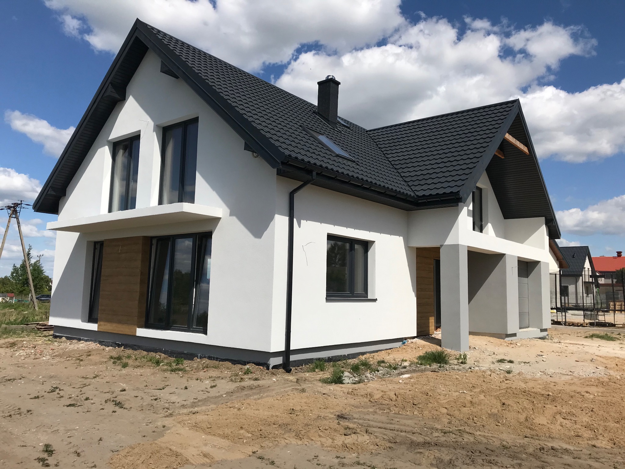 Nowoczesny, parterowy budynek z białą elewacją, ciemną dachówką i drewnianymi akcentami, usytuowany na działce w trakcie zagospodarowywania.