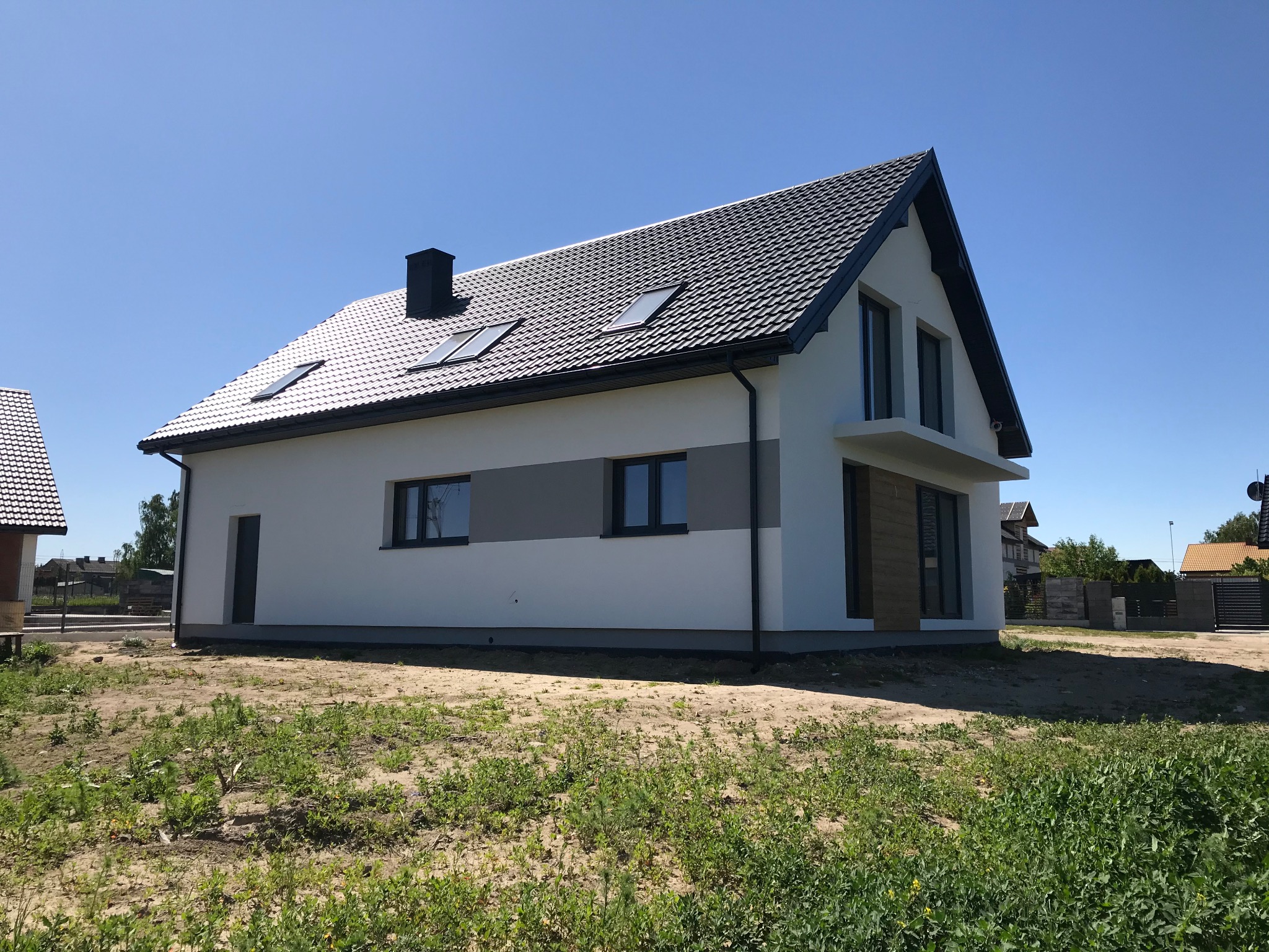 Nowoczesny, dwukondygnacyjny dom z ciemną dachówką i białymi ścianami, z szarym pasem dekoracyjnym, widoczny z zewnątrz na tle błękitnego nieba i niezagospodarowanego terenu z zieloną roślinnością.