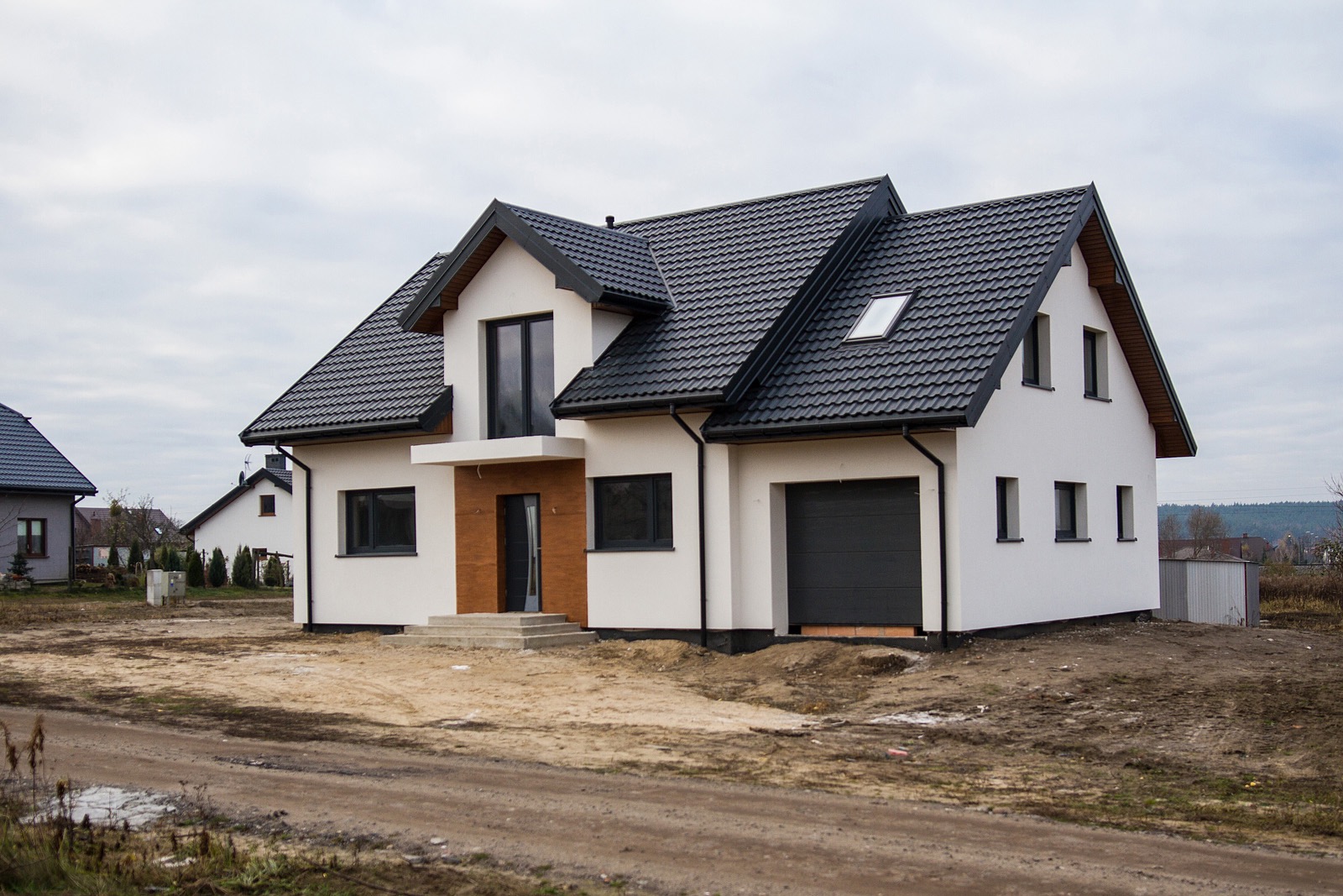 Nowoczesny dom murowany z czarną dachówką i garażem, surowy teren wokół budynku, widoczne inne domy w tle.