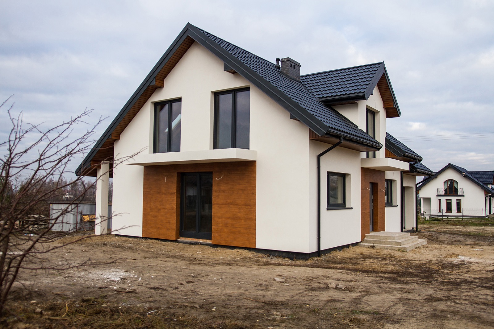 Nowoczesny dom z białymi ścianami, ciemną dachówką i elementami dekoracyjnymi z paneli imitujących drewno, widoczny w pochmurny dzień, z drugim domem w tle.