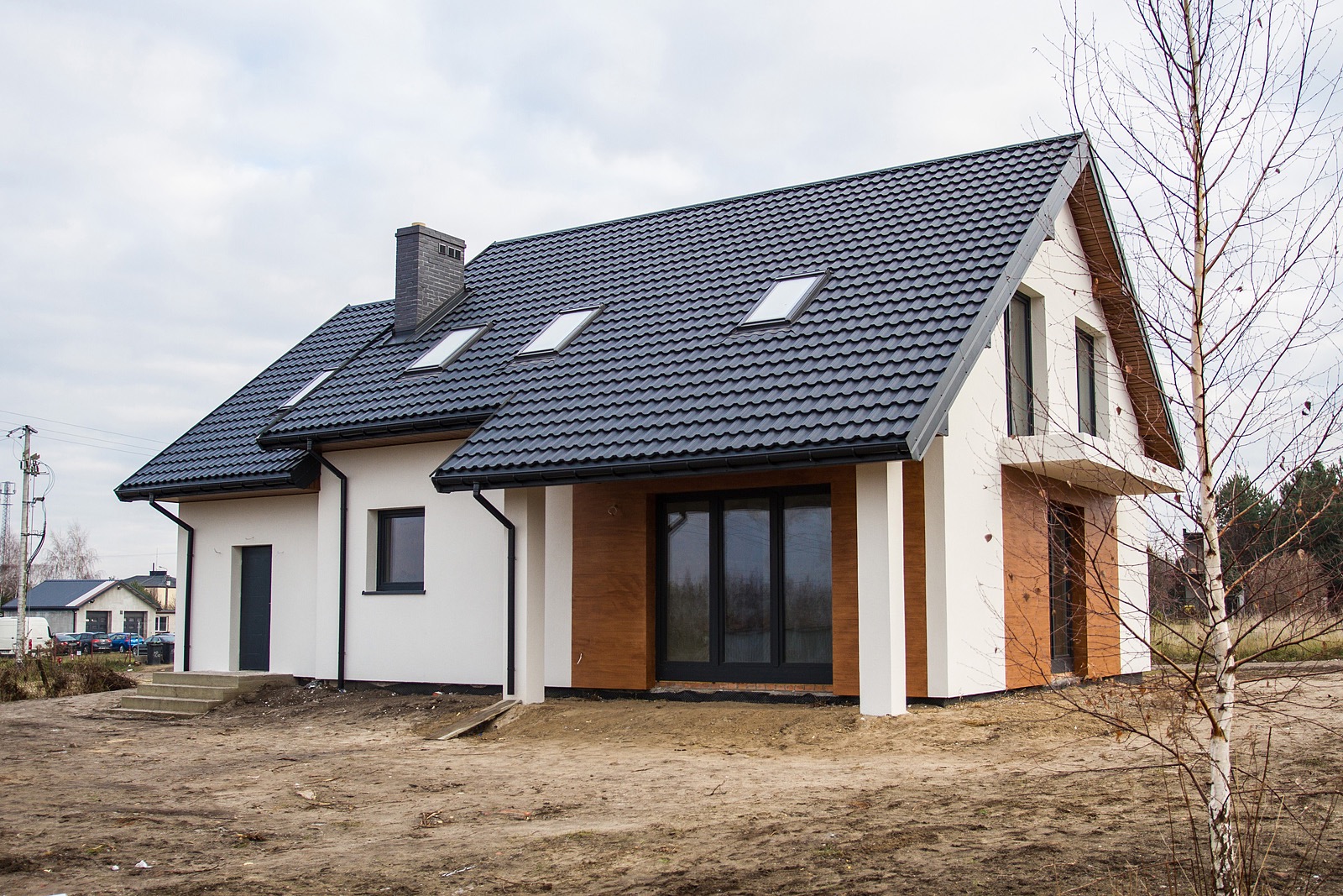 Nowoczesny, parterowy budynek z ciemną, skośną dachówką, trzema oknami dachowymi i wykończeniem elewacji w bieli i imitacji drewna, widoczny z zewnątrz w pochmurny dzień.