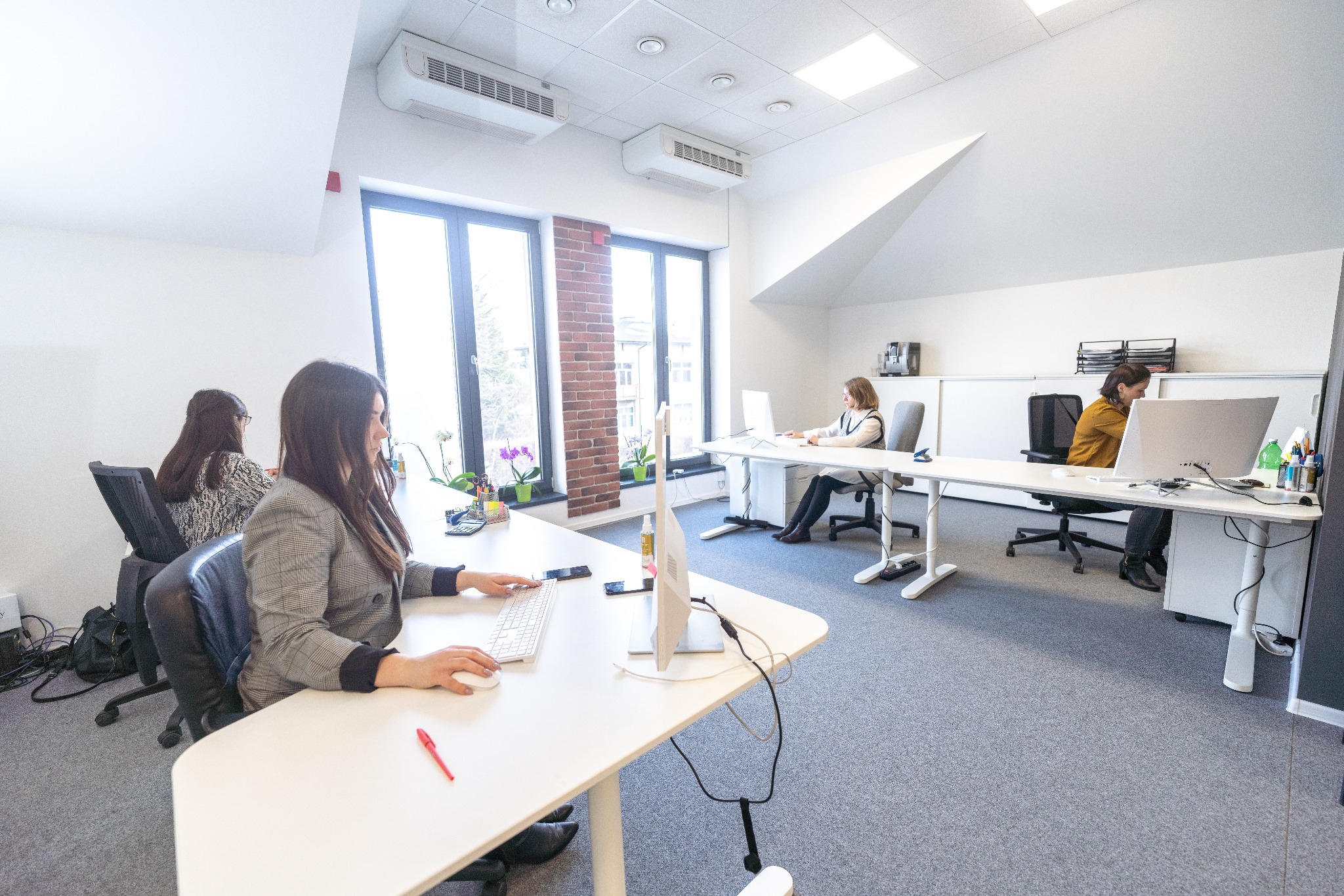 Jasne biuro z pracownikami przy biurkach, kobieta w kraciastej marynarce pisze na klawiaturze, na biurku czerwony długopis, okno z widokiem na ceglany budynek.