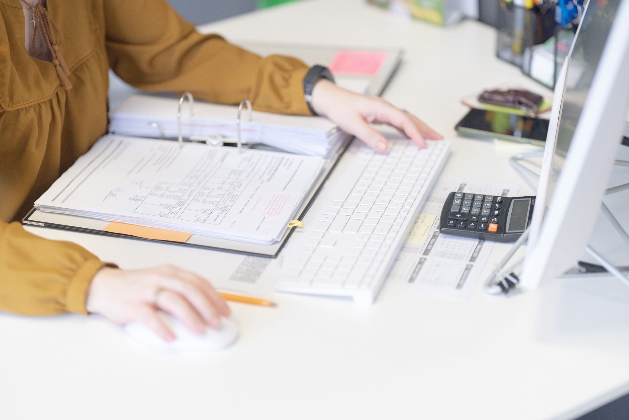 Kobieta w musztardowej bluzce pracuje przy biurku z dokumentami w segregatorze, klawiaturą, kalkulatorem i kalendarzem, używając myszki komputerowej.