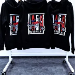 Trzy czarne bluzy z kapturem wiszące na wieszakach, z nadrukiem czerwonego motocykla i napisem 'JACK MOTORS.PL' na plecach, na tle białej ściany i szarej podłogi.