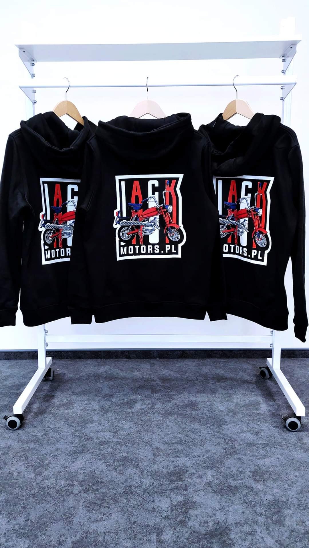 Trzy czarne bluzy z kapturem wiszące na wieszakach, z nadrukiem czerwonego motocykla i napisem 'JACK MOTORS.PL' na plecach, na tle białej ściany i szarej podłogi.