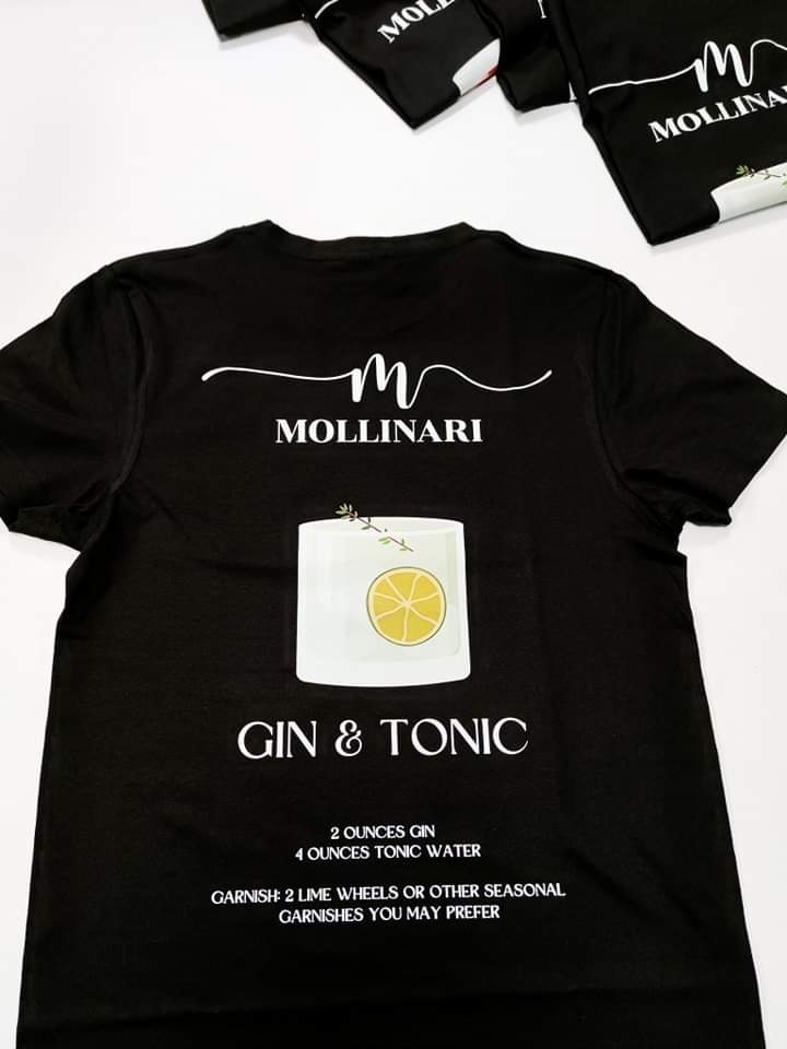 Czarna koszulka z nadrukiem Gin & Tonic, logo firmy Mollinari oraz przepisem na drinka, ułożona na białym tle obok innych koszulek z tym samym nadrukiem.