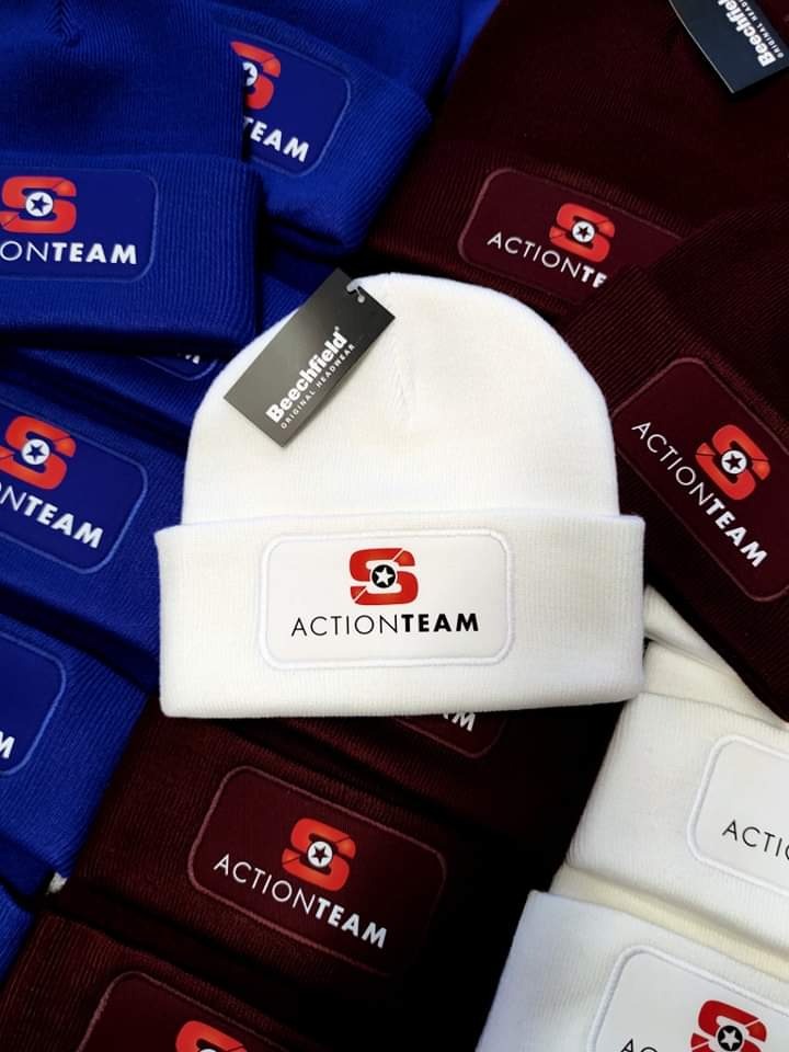 Czapki zimowe w kolorach niebieskim, białym i bordowym, każda z naszytym logo 'ACTIONTEAM' na prostokątnej naszywce, biała czapka z metką 'Beechfield'.