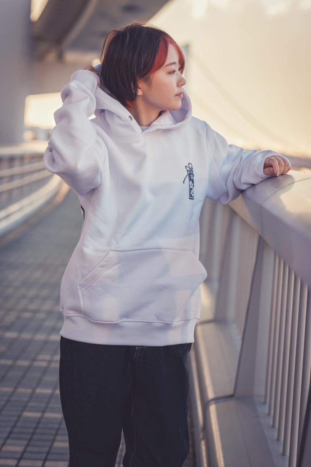 Młoda kobieta w białej bluzie z kapturem i ciemnych spodniach, oparta o barierkę na tle zachodzącego słońca. Na bluzie widoczny mały, ciemny nadruk.