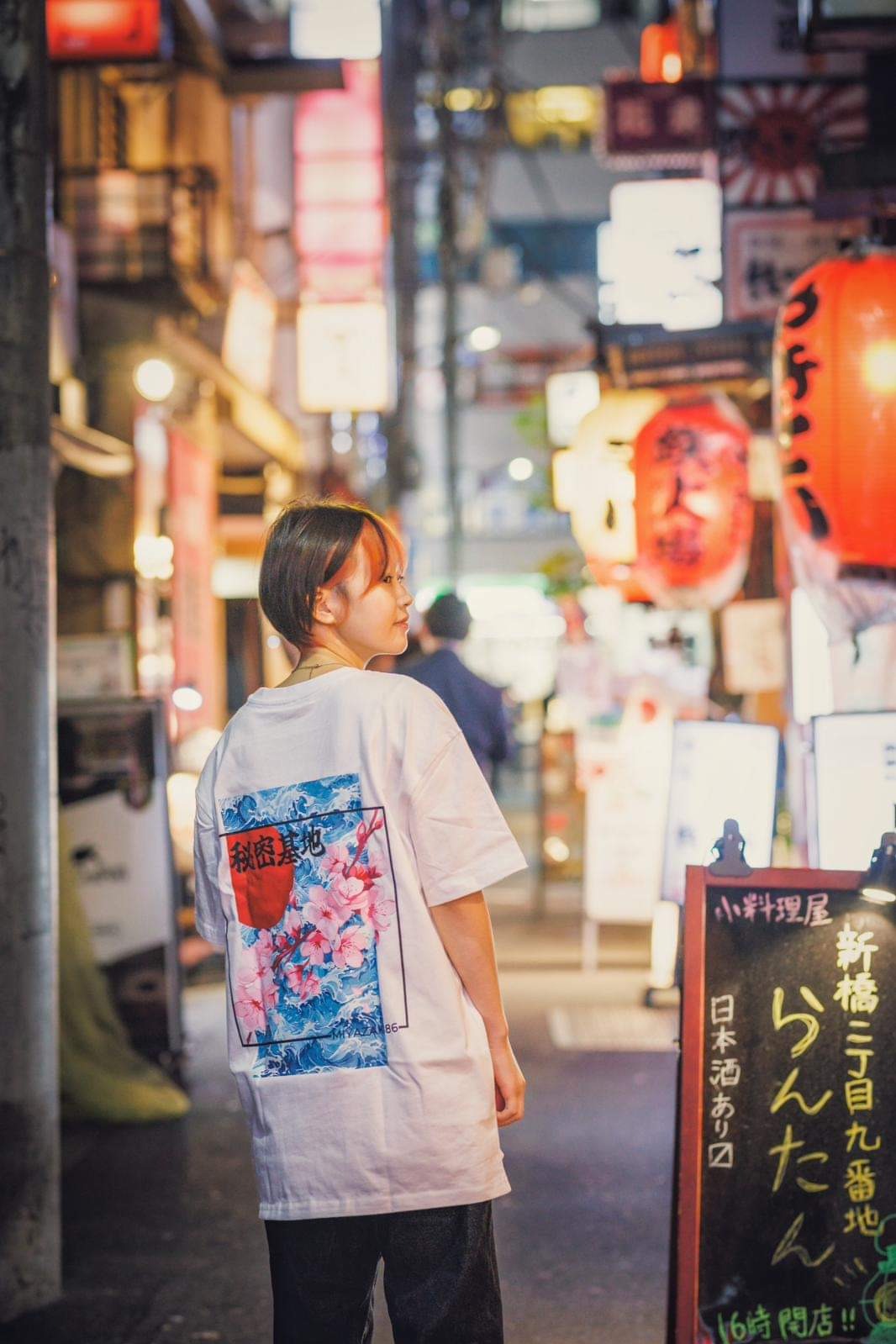 Dziewczyna w białej koszulce z nadrukiem kwiatów i fal w stylu japońskim, stojąca tyłem na ulicy w Tarnowie, w tle lampiony i szyldy reklamowe.