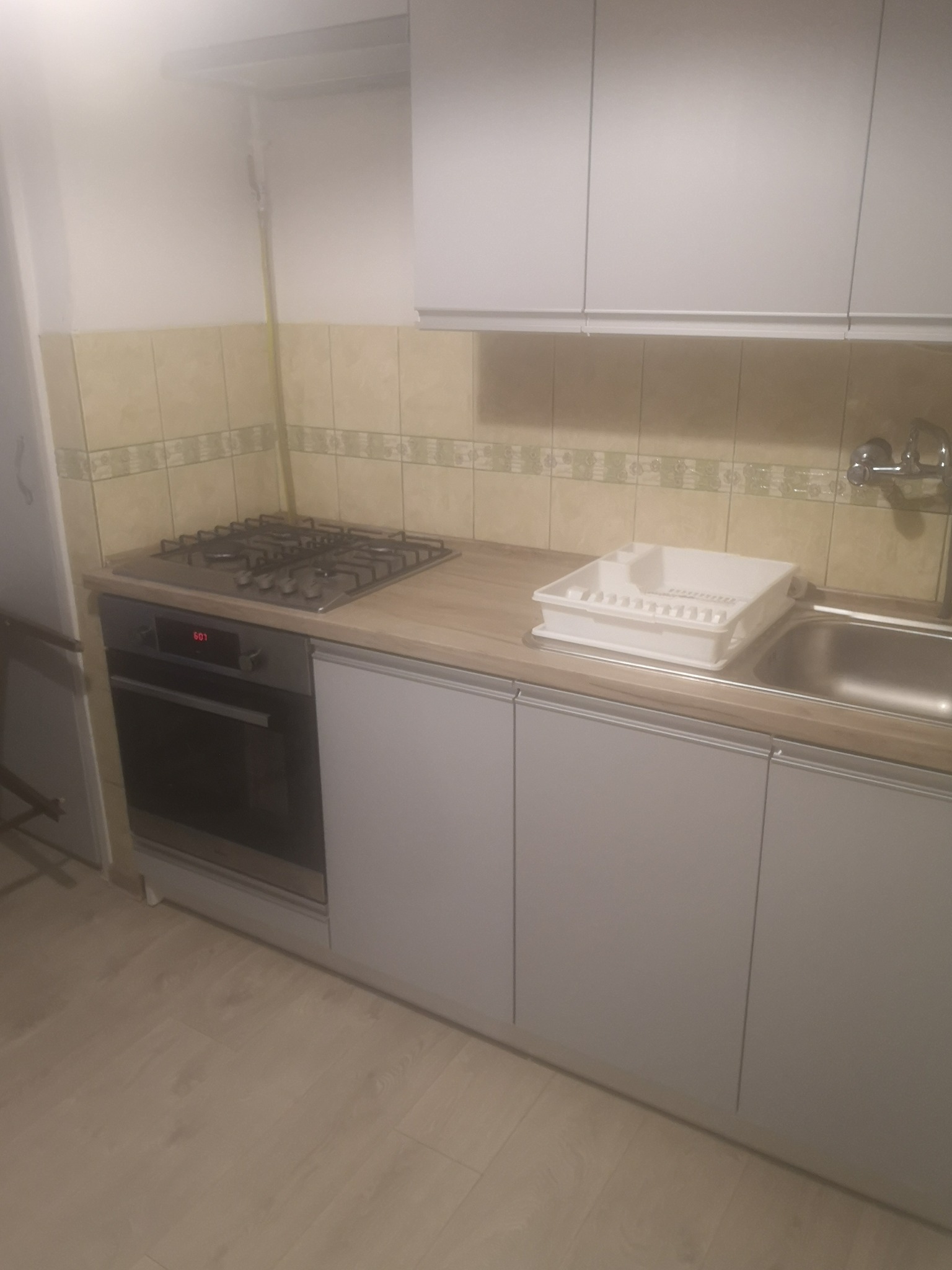 Kuchnia z białymi szafkami, beżowym blatem, płytą gazową i zlewozmywakiem ze suszarką do naczyń, widok na fragment ściany z beżowymi płytkami.