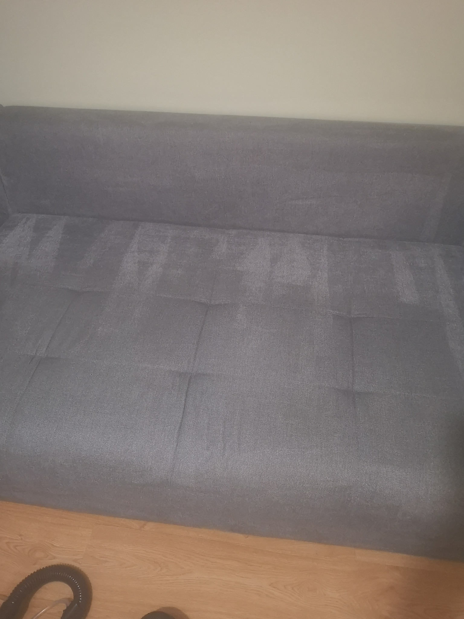 Szara kanapa z widocznymi śladami czyszczenia, jaśniejsze fragmenty na ciemniejszym tle, fragment węża odkurzacza widoczny na podłodze.