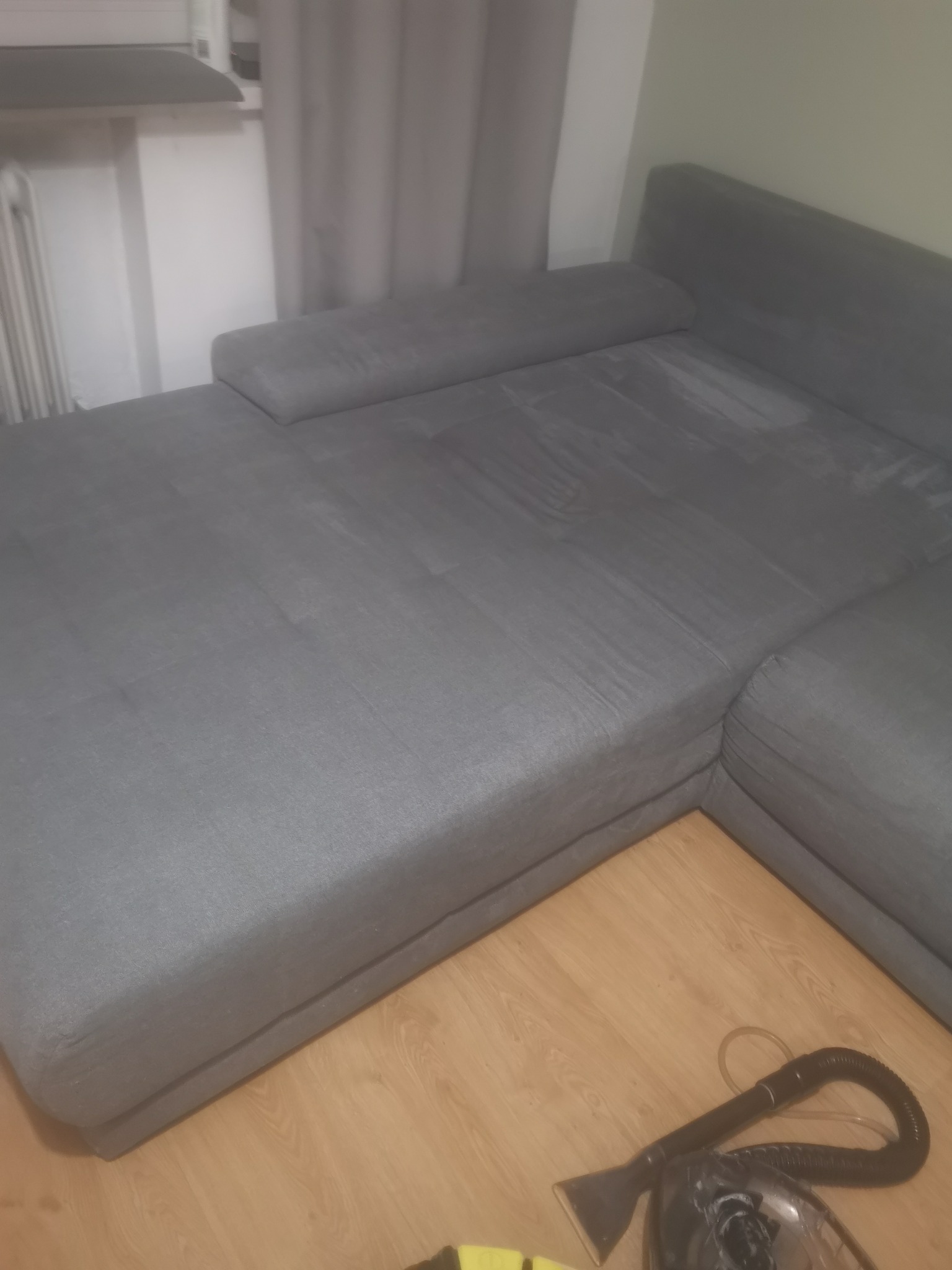 Szara kanapa narożna z widocznymi śladami czyszczenia i urządzeniem do prania tapicerki leżącym na podłodze.