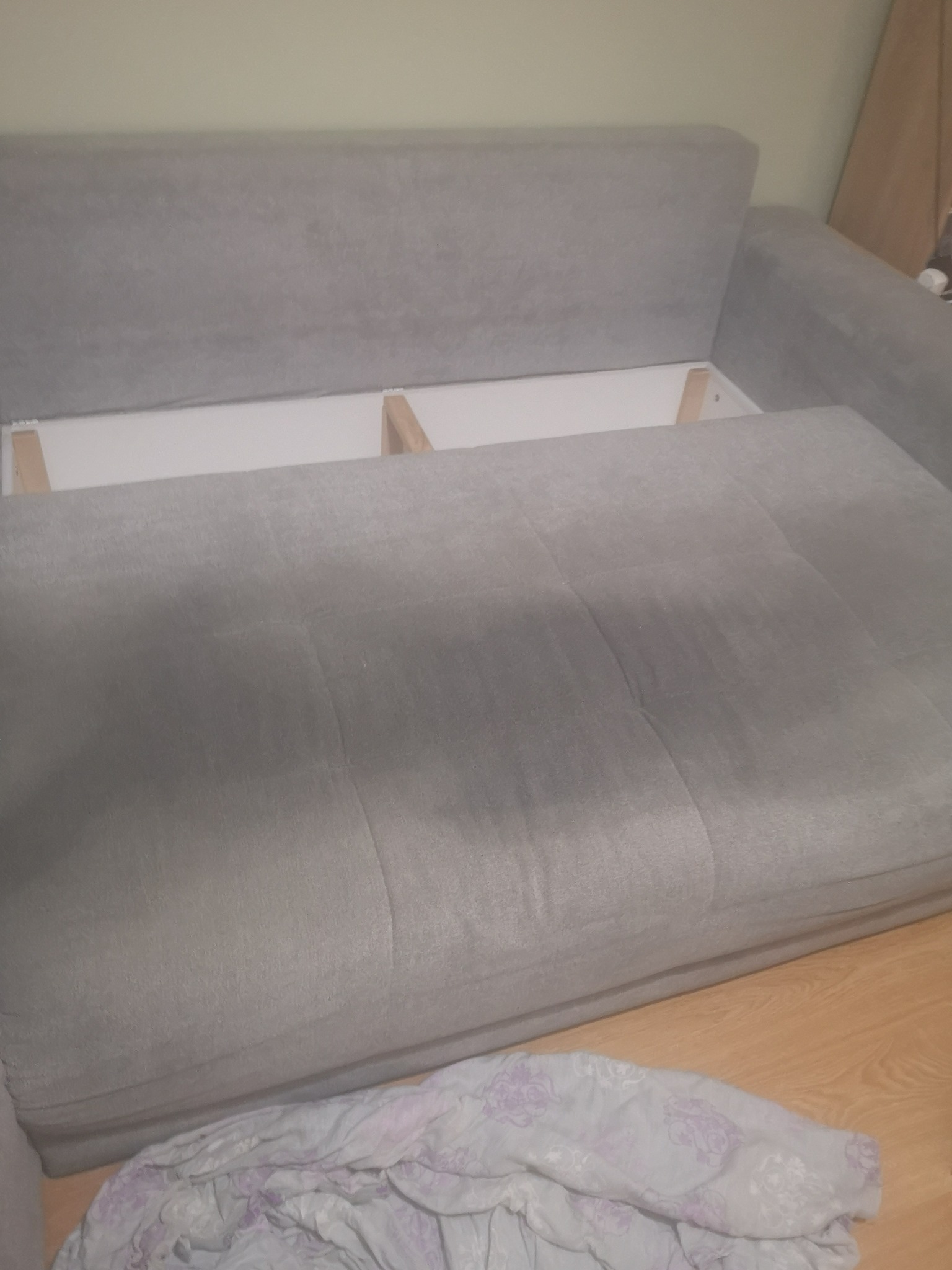 Szara kanapa z podniesionym siedziskiem, ukazującym biały pojemnik na pościel z drewnianymi wzmocnieniami, na podłodze leży zmięta pościel w kwiaty.