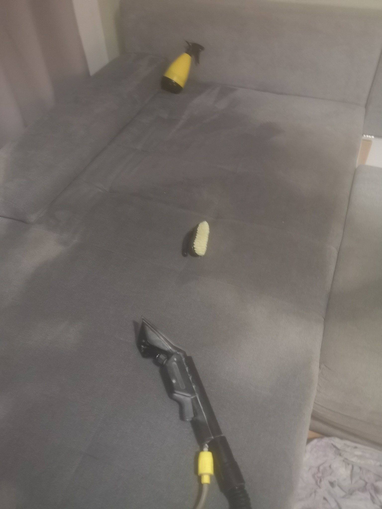 Szara kanapa z widocznymi plamami, w trakcie czyszczenia: odkurzacz piorący z dyszą, żółty spryskiwacz i szczotka do tapicerki.