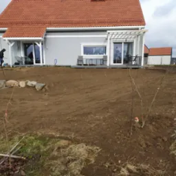 Przygotowany teren pod ogród z młodymi drzewkami owocowymi w pobliżu nowoczesnego domu z czerwonym dachem, widoczne narzędzia ogrodnicze i pracownik w tle.