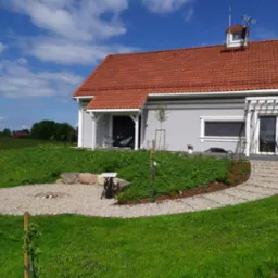 Nowoczesny dom z ogrodem: trawnik, ścieżka z kamienia prowadząca do domu, okrągłe miejsce na ognisko z kamieniami, młode drzewo podparte palikiem, rabata z roślinami i korą, oraz dekoracyjny stolik...