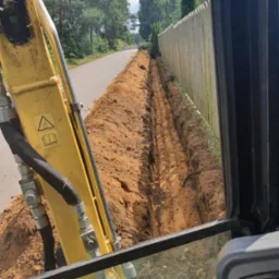 Widok z kabiny koparki na wykopany rów wzdłuż drewnianego płotu i asfaltowej drogi, z widocznymi śladami gąsienic na dnie wykopu.