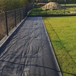 Water Garden - Przygotowanie terenu pod trawnik z rolki: czarna mata rozłożona na ziemi, obok zielona trawa i ogrodzenie panelowe. Widok na domy w tle.