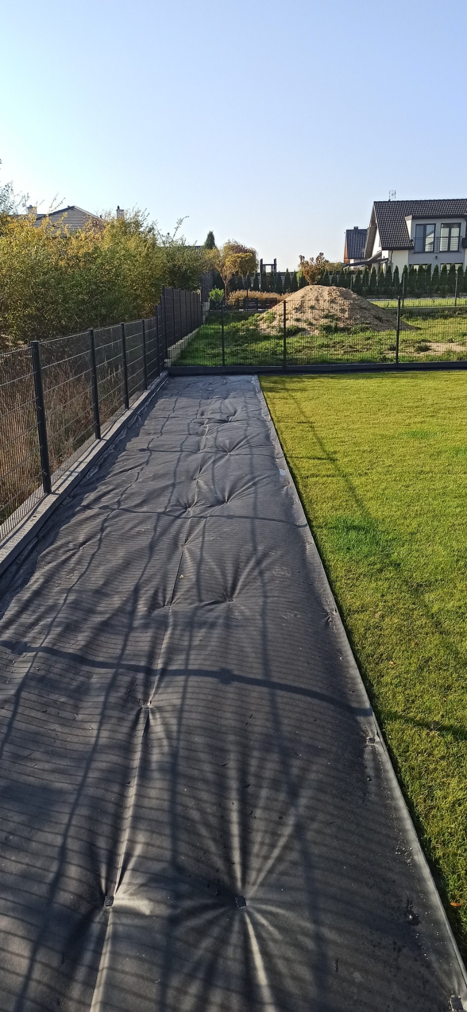 Przygotowanie terenu pod trawnik z rolki: czarna mata rozłożona na ziemi, obok zielona trawa i ogrodzenie panelowe. Widok na domy w tle.