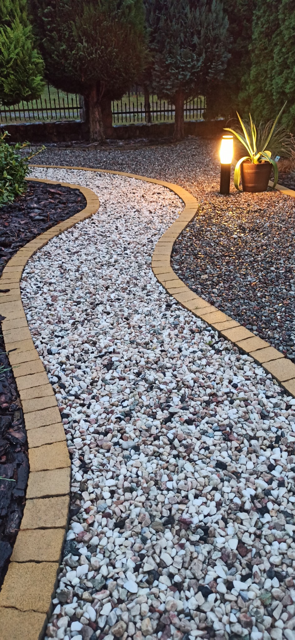 Kręta ścieżka z białego żwiru, obrzeża z żółtej kostki, oświetlona lampą ogrodową. Tło z ciemnej kory i zieleni. Widok na zadbany ogród.