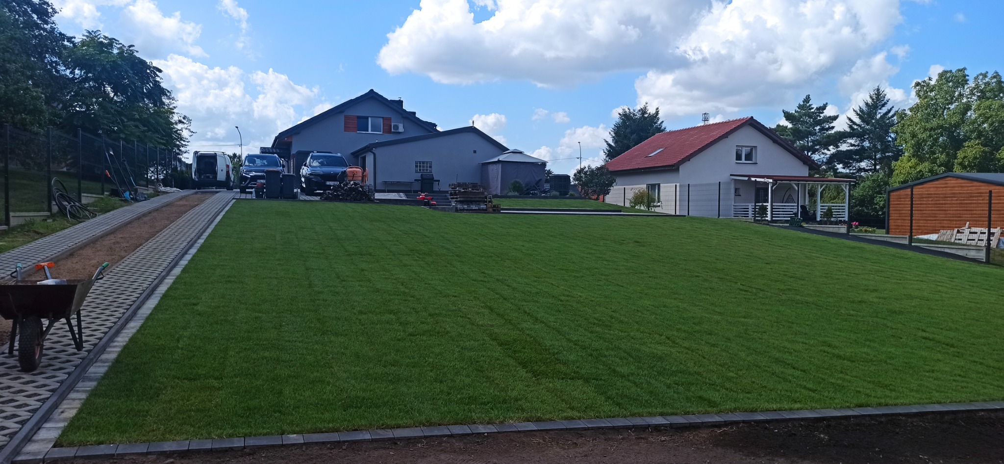 Równy trawnik z rolki na posesji z dwoma domami, w tle zaparkowane samochody i narzędzia ogrodnicze. Widoczne obrzeża trawnika i kostka brukowa.