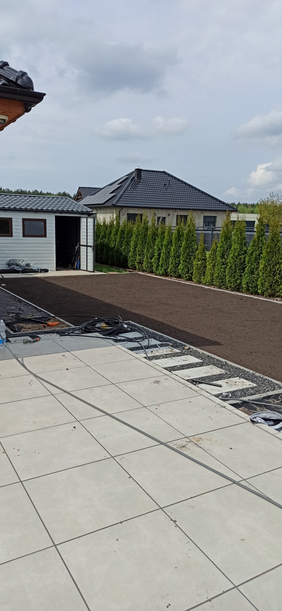 Przygotowany teren pod trawnik z rolki, obrzeża z kamienia, widok z tarasu w trakcie budowy. W tle rząd tuj i budynek z panelami słonecznymi.