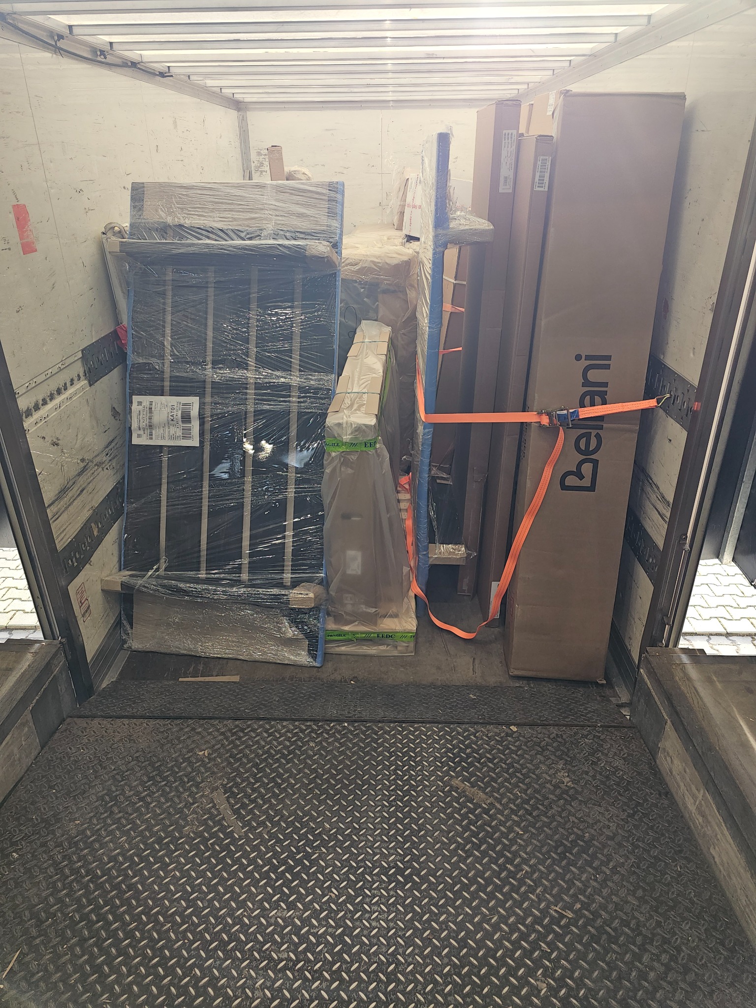 Wnętrze furgonetki wypełnione po brzegi meblami i kartonami zabezpieczonymi pomarańczowymi pasami transportowymi, widoczna duża paczka z logo Bellani.