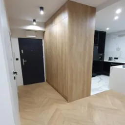 Wysoka szafa z drewnopodobnymi frontami, oddzielająca przedpokój od kuchni z czarnymi meblami i marmurową podłogą, parkiet ułożony w jodełkę.