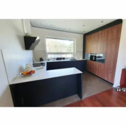 Nowoczesna kuchnia z wyspą, czarne szafki dolne z białym blatem, drewniane szafki górne, okno z widokiem na zieleń, czarny okap i miska z owocami na blacie.