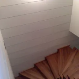 Drewniane schody spiralne z widoczną ścianą wykończoną panelami w odcieniach szarości, ujęcie z góry.