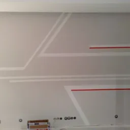 Ściana w jasnych odcieniach szarości z geometrycznymi wzorami w kolorze białym i czerwonym, widoczne gniazdka elektryczne i puszki podtynkowe, kartonowe pudełko na podłodze.