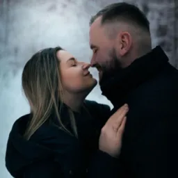 Para w zimowej scenerii, zbliżenie twarzy, dotykające się nosami, kobieta z pierścionkiem na palcu, oboje w ciemnych kurtkach na tle ośnieżonego lasu.