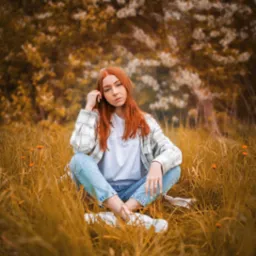 Portret młodej kobiety z rudymi włosami siedzącej w trawie na tle kwitnącego drzewa, ubrana w jeansy, białą koszulkę i flanelową koszulę w kratę.