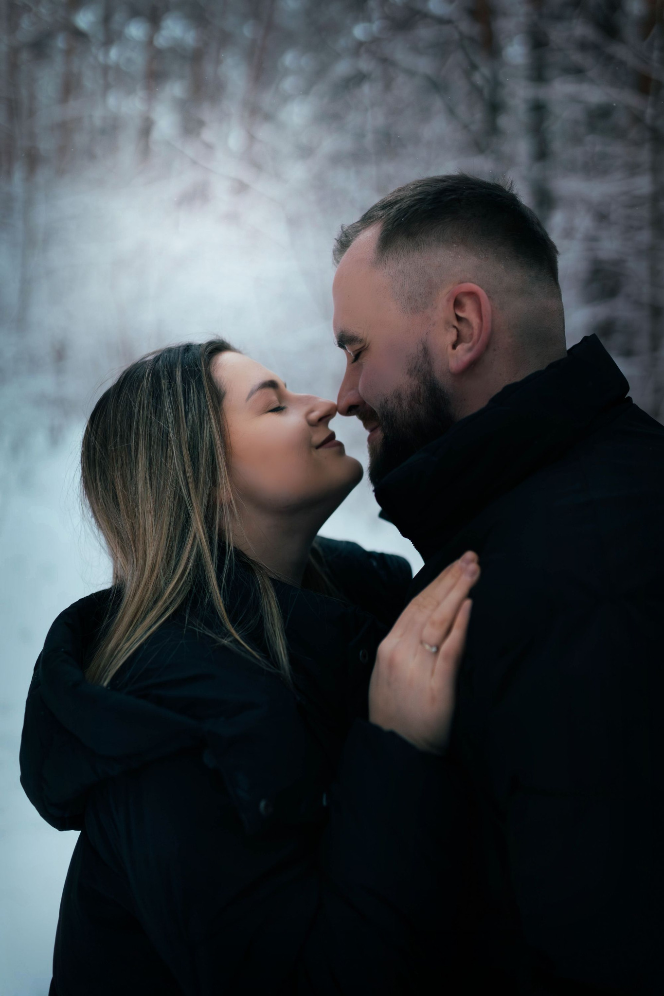 Para w zimowej scenerii, zbliżenie twarzy, dotykające się nosami, kobieta z pierścionkiem na palcu, oboje w ciemnych kurtkach na tle ośnieżonego lasu.