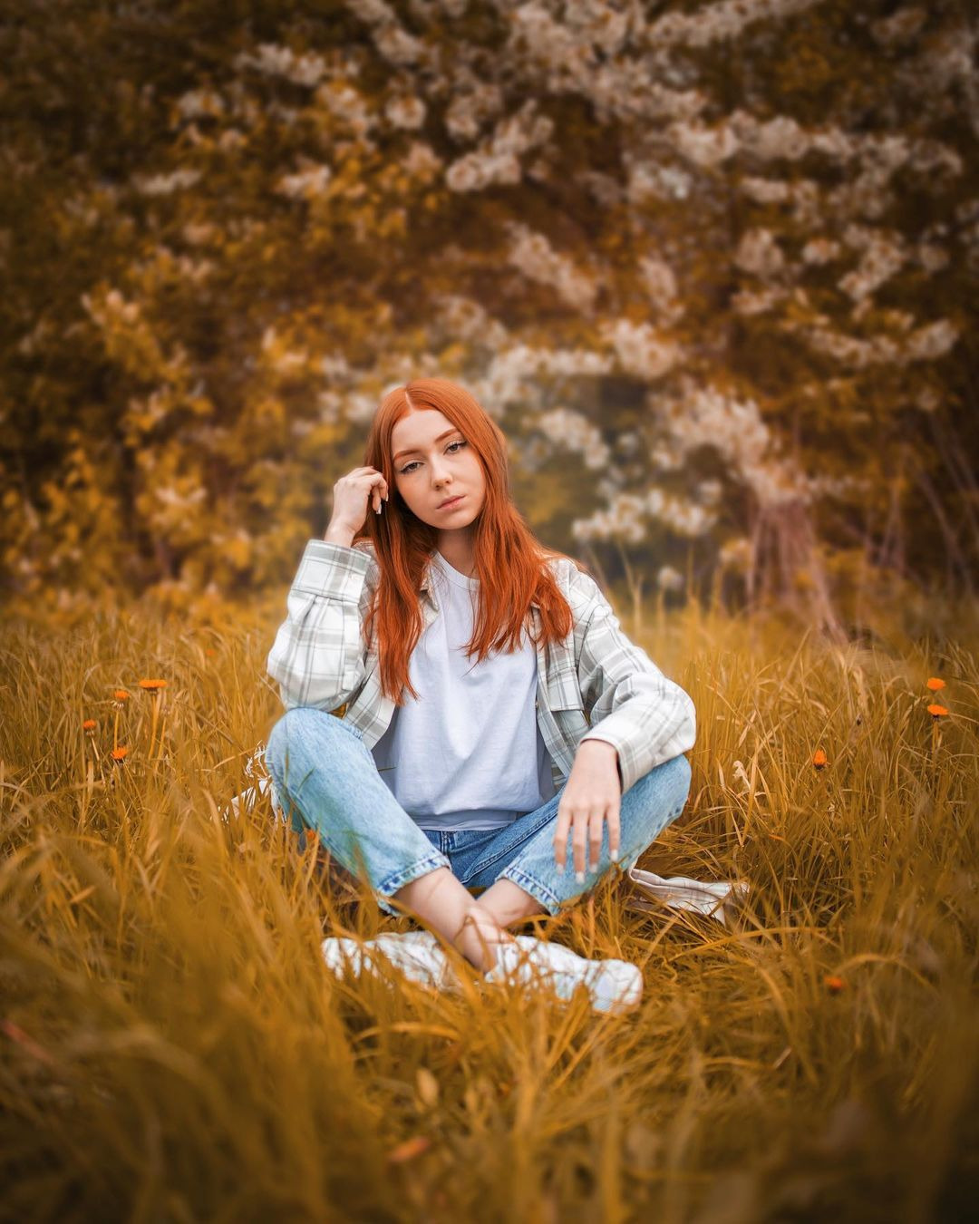 Portret młodej kobiety z rudymi włosami siedzącej w trawie na tle kwitnącego drzewa, ubrana w jeansy, białą koszulkę i flanelową koszulę w kratę.