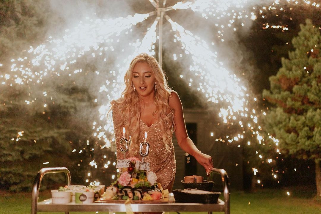 Kobieta w złotej sukience kroi tort urodzinowy z kwiatami i sushi, na tle fontanny iskier.