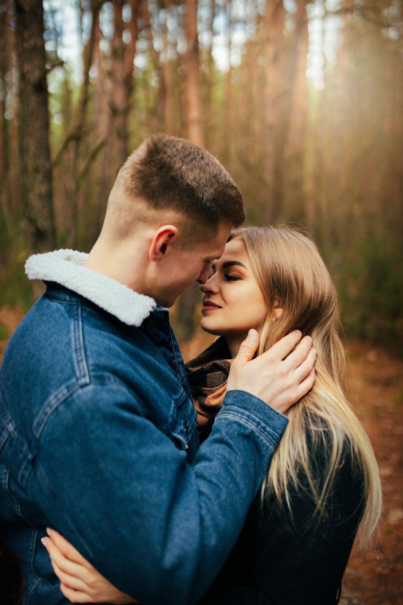 Para zakochanych w lesie, mężczyzna w dżinsowej kurtce z barankiem obejmuje kobietę, która delikatnie dotyka jego twarzy; ciepłe światło słoneczne w tle.